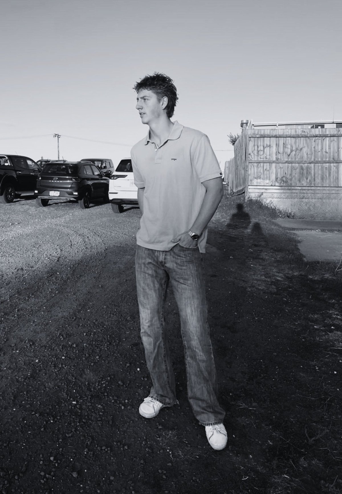 Young man standing in a junkyard wearing a polo shirt and jeans, looking to the side with parked cars and a metal gate in the background